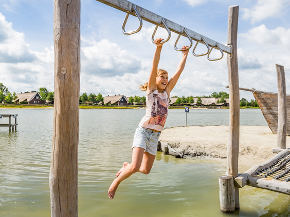 Een meisje slingert aan het klimrek met ringen boven het water van het recreatiemeer op het speeleiland van Landal Hof van Saksen.