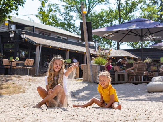 Twee kinderen spelen vrolijk in het witte zand op het strandterras van het restaurant bij Landal Klein Oisterwijk in Noord-Brabant.