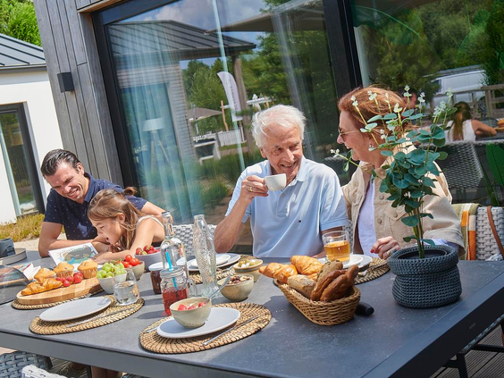 Een familie met opa, oma, vader en dochter geniet van een uitgebreid ontbijt met croissantjes en jus d'orange op het zonnige terras van hun vakantiehuis op Landal Klein Oisterwijk.