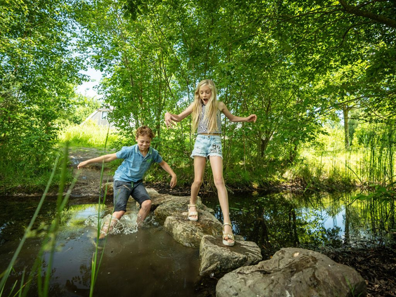 Een broer en zus balanceren op grote stenen in een waterplas, omringd door het groen van de natuurspeeltuin op Landal Klein Oisterwijk.