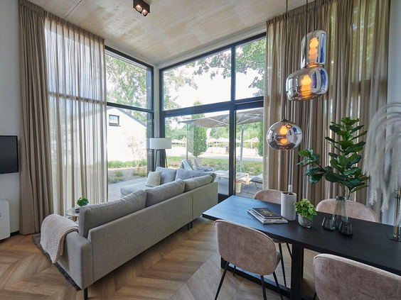 Luxe en moderne woonkamer in een vakantiehuis op Landal Klein Oisterwijk, met extra hoge ramen die een prachtig uitzicht bieden op de tuin en veel natuurlijk licht binnenlaten.