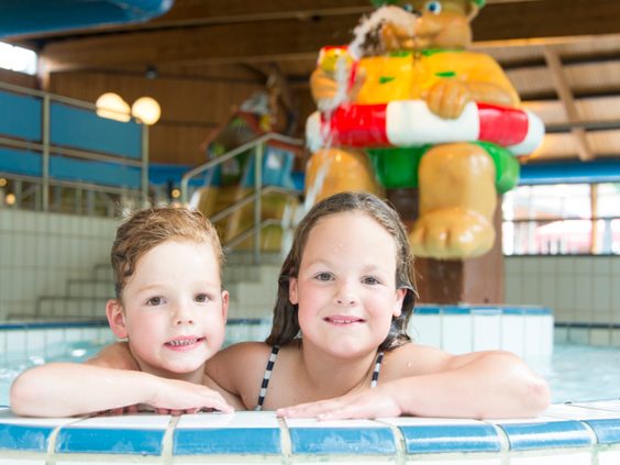 Twee blije kinderen poseren in het binnenzwembad van Landal Miggelenberg, met een beeld van Bollo de beer op de achtergrond.