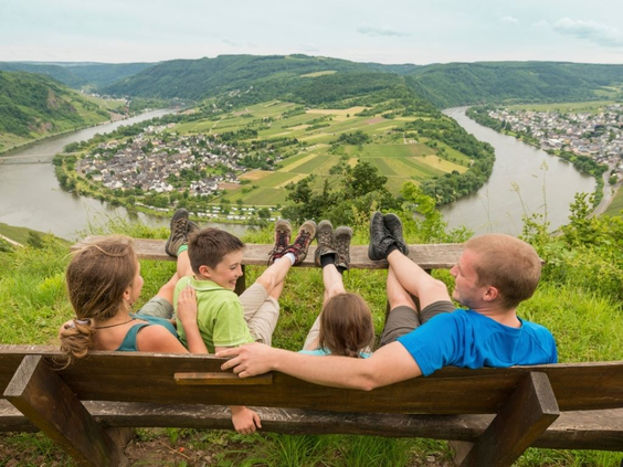 Een gezin rust uit op een bankje en geniet van het panoramische uitzicht over de beroemde bocht in de rivier de Moezel, na een wandeling vanuit Landal Mont Royal.