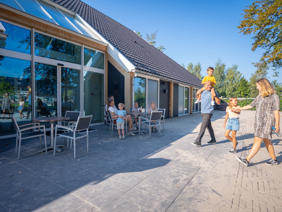 Gezin wandelt langs het zonnige terras van het restaurant bij het centrumgebouw op Landal Orveltermarke.