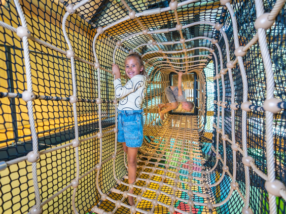 Meisje klimt lachend door een lange, avontuurlijke touwtunnel in het indoor speelparadijs van Landal Orveltermarke.