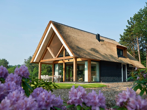 Een luxe vakantieboerderij met rieten dak en grote ramen op Landal Puur Exloo, omringd door bloeiende paarse bloemen op een zonnige dag.