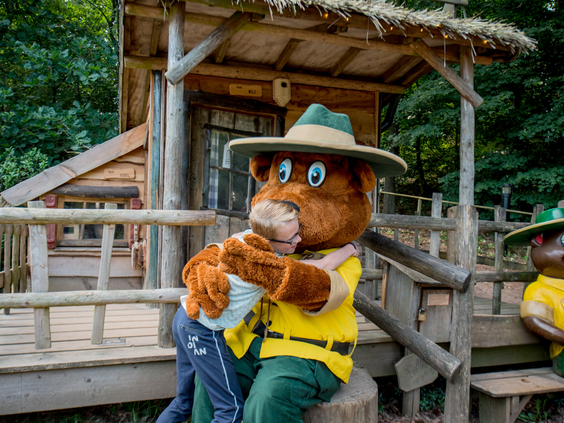 Een kind geeft een dikke knuffel aan mascotte Bollo de Beer, die bij een houten speelhuisje zit op vakantiepark Landal Rabbit Hill op de Veluwe.