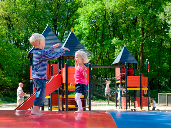 Twee jonge, blije kinderen springen op een grote rode airtrampoline in de buitenspeeltuin van Landal Rabbit Hill, met op de achtergrond een houten speelkasteel.