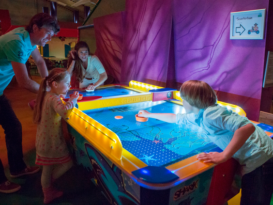 Kinderen en volwassenen spelen een fanatiek potje airhockey aan een kleurrijke, verlichte speeltafel in de indoor spelhal van Landal Rabbit Hill.