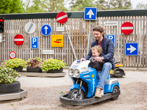 Een vader en zoon rijden samen op een blauwe speelgoedscooter door het verkeerspark met verkeersborden op vakantiepark Landal Rabbit Hill.