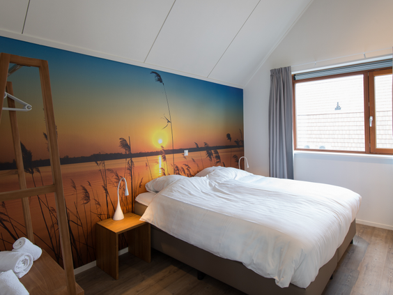 Sfeervolle slaapkamer in een waterwoning op Landal Reeuwijkse Plassen, met een comfortabel opgemaakt bed en een opvallende fotowand van een zonsondergang over het water.