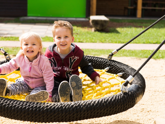 Twee lachende peuters zitten samen in de grote nestschommel in de buitenspeeltuin van vakantiepark Landal Salztal Paradies.