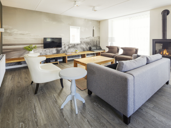 Een stijlvol ingerichte, moderne woonkamer met een grijze hoekbank, een houtkachel en een grote fotowand van het Texelse strand in een bungalow op Landal Sluftervallei.