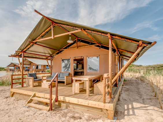 De achterzijde van een stoere safaritent met een houten veranda, gelegen in het zanderige duinlandschap van vakantiepark Landal Sluftervallei op Texel.