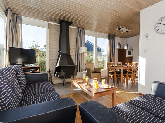 Een gezellig ingerichte woonkamer van een bungalow op Landal Sluftervallei, met blauwe banken, een houtkachel en een eethoek bij het raam.