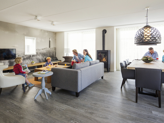 Een grote, open woonkamer van een groepsaccommodatie op Landal Sluftervallei, waar een familie ontspant op de banken en aan de lange eettafel.