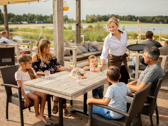 Gezin met kinderen geniet van een maaltijd op het buitenterras van het restaurant aan het water bij Landal Stroombroek.