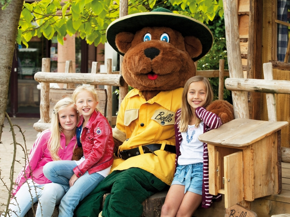 Drie vrolijke meisjes zitten op een houten bankje en poseren voor een foto met de mascotte Bollo de Beer.