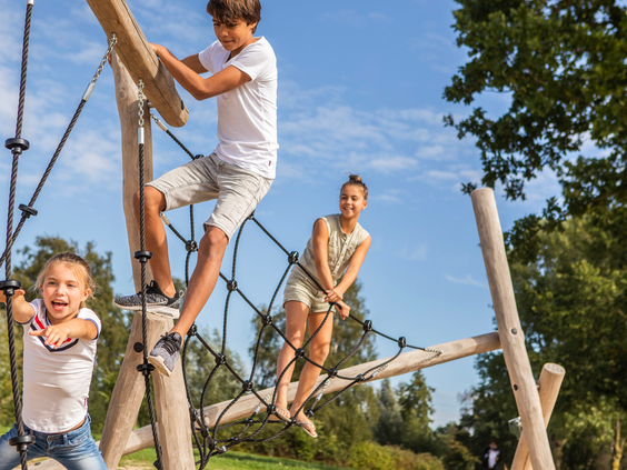 Drie kinderen van verschillende leeftijden klimmen en balanceren op een avontuurlijk klimrek van hout en touwen.