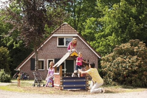 Kinderen spelen met hun ouders bij een speeltoestel met glijbaan in de tuin van een kinderbungalow op Landal Stroombroek.