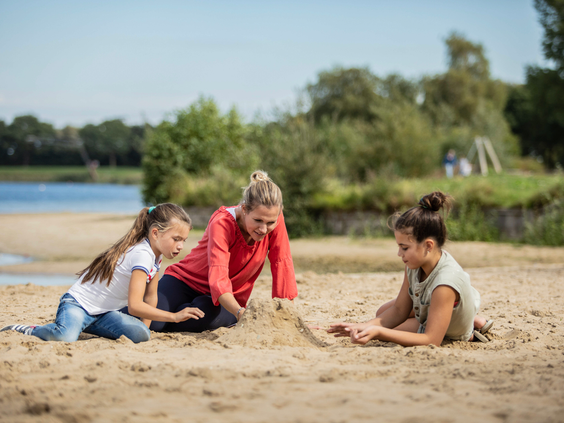 Moeder bouwt met haar twee dochters een zandkasteel op het zandstrand van de recreatieplas bij Landal Stroombroek.