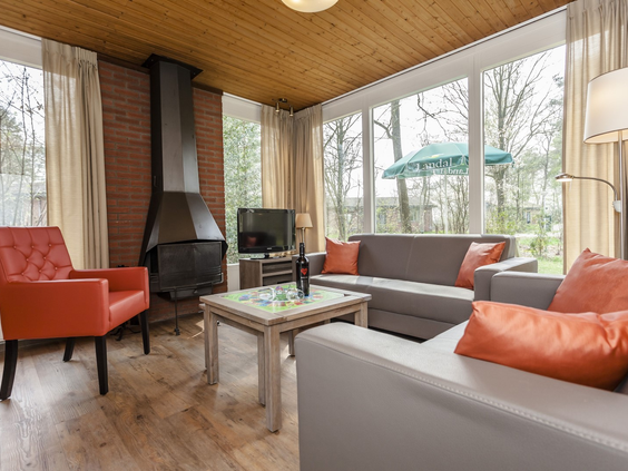 Een gezellige woonkamer met een grote open haard, comfortabele bank, oranje fauteuil en uitzicht op de tuin bij Landal Twenhaarsveld.