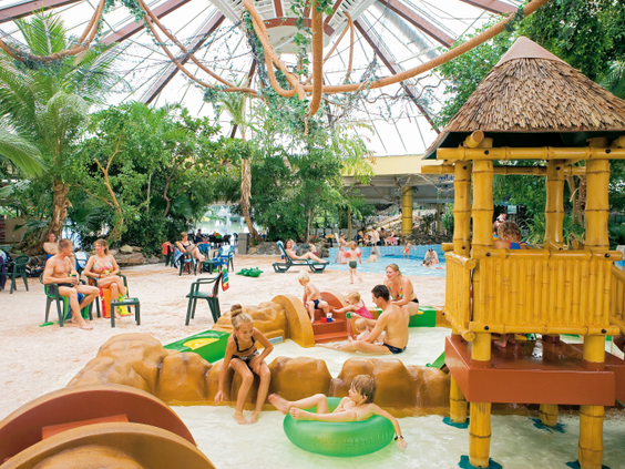 Kinderen spelen op en rond een houten speeltoestel in het ondiepe, veilige kinderbad van het subtropisch zwemparadijs op Landal Vennenbos.