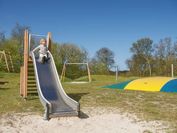 Een kind glijdt van de glijbaan in de buitenspeeltuin van Landal Vitamaris, met op de achtergrond een grote airtrampoline.