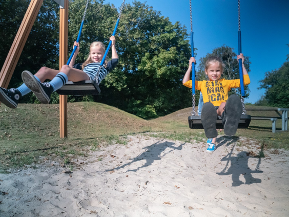Twee blije kinderen schommelen hoog in de lucht in de zanderige speeltuin van Landal Vitamaris op Schiermonnikoog.