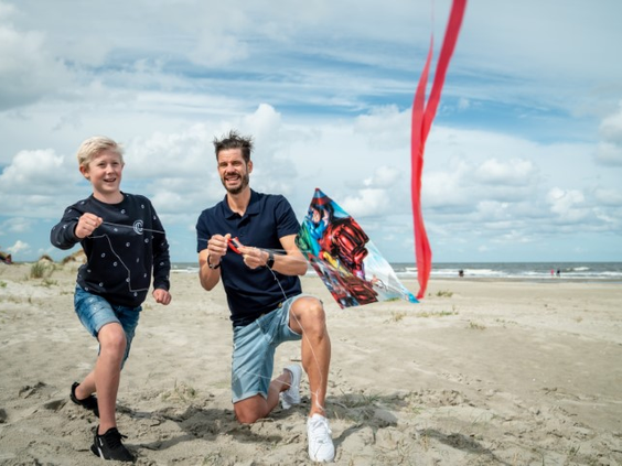 Een vader en zoon rennen enthousiast over het strand van Schiermonnikoog terwijl ze samen een vlieger oplaten.