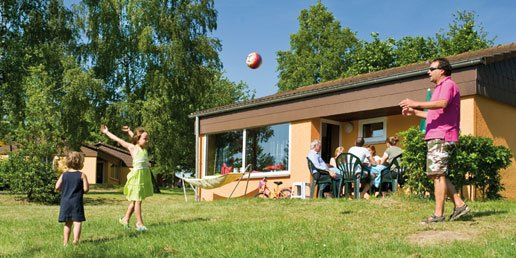 Een meisje gooit een bal in de lucht op het grasveld voor de bungalow op Landal Warsberg, terwijl haar familie op het terras zit.
