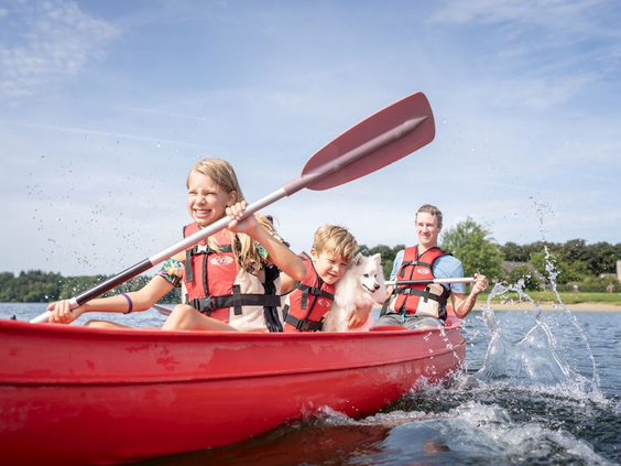 Een lachend gezin met twee kinderen en zwemvesten aan, peddelt in een rode kano op het water bij Landal Waterresort Blocksyl.