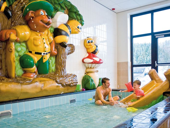 Een vader speelt met zijn kind in het ondiepe peuterbad van het zwembad op Landal Winterberg, compleet met speeltoestellen en een kleine glijbaan.