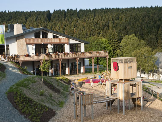 Het hoofdgebouw van Landal Winterberg met receptie en restaurant, met op de voorgrond een grote houten speeltuin in de buitenlucht.