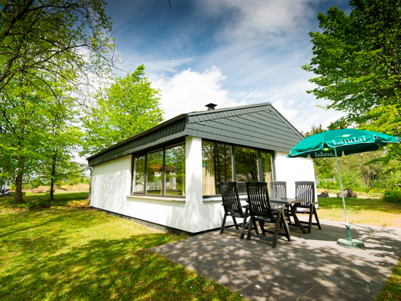 Een vrijstaande, witte bungalow van Landal Wirfttal met een terras en tuinmeubilair, gelegen in een groene, bosrijke omgeving.