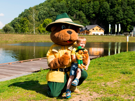Bollo de Beer, de mascotte van Landal, geeft een high five aan een blij kind aan de oever van het meer op vakantiepark Wirfttal.