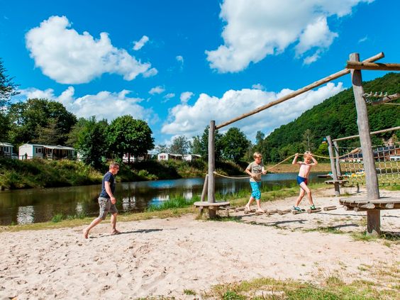 Kinderen spelen en balanceren op houten speeltoestellen op het zandstrand aan het recreatiemeer van Landal Wirfttal.