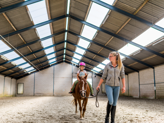 Een meisje met een cap op krijgt paardrijles op een pony in de overdekte manege van Landgoed de Scheleberg op de Veluwe.