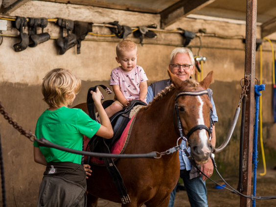 Een klein kind zit trots op de rug van een pony in de stal van de manege op Landgoed de Scheleberg, terwijl het wordt vastgehouden.