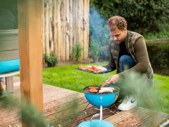 Een man is aan het barbecueën op een kleine barbecue in de tuin van een vakantiehuisje op Landgoed de Scheleberg.