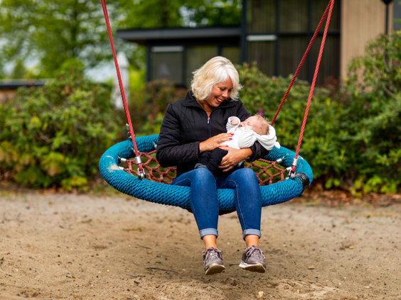 Een oma zit met een slapende baby op haar schoot zachtjes te wiegen op de grote nestschommel in de speeltuin.