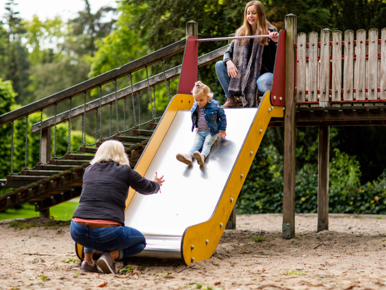 Een kind glijdt van de glijbaan in de speeltuin, terwijl moeder en oma toekijken en helpen op Landgoed de Scheleberg.