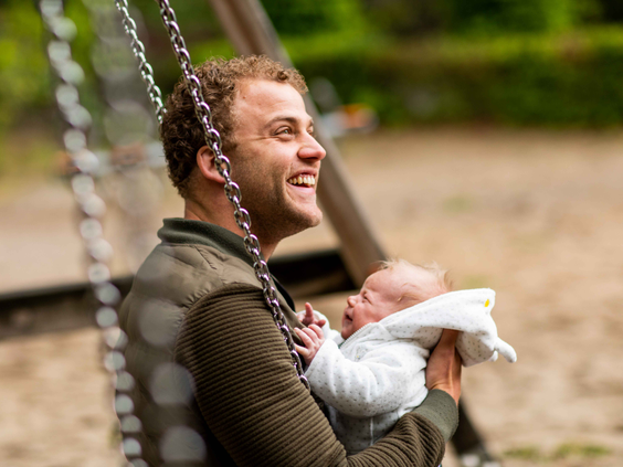 Een lachende vader zit met zijn pasgeboren baby op schoot op een schommel in de speeltuin van Landgoed de Scheleberg.