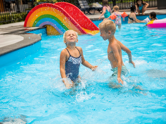 Twee kinderen spelen vrolijk in het ondiepe peuterbad met op de achtergrond een kleurrijke regenboogglijbaan.
