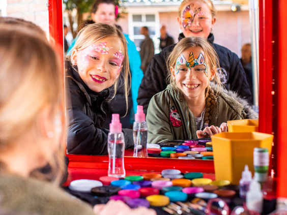 Twee lachende meisjes kijken in de spiegel naar hun gezicht dat wordt geschminkt met glitters tijdens een activiteit van het animatieteam.