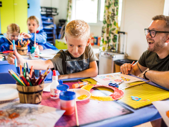 Kinderen zitten met een vader aan een tafel vol met potloden en verf te knutselen tijdens een activiteit van de kidsclub.