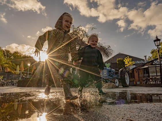 Twee kinderen springen en stampen vrolijk in een grote waterplas op Landgoed Ruwinkel, met de zon op de achtergrond.