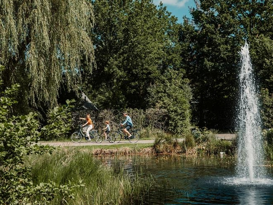 Een gezin fietst langs een grote vijver met een fontein op een zonnige dag op Landgoed Ruwinkel.