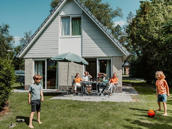 Familie ontspant op het terras in de tuin van hun vakantiehuis, terwijl de kinderen aan het voetballen zijn op het gras.