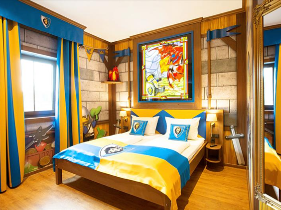 Thematische ridderhotelkamer in het LEGOLAND Feriendorf, met een groot bed, blauw-gele dekens en middeleeuwse decoraties voor een unieke overnachting.